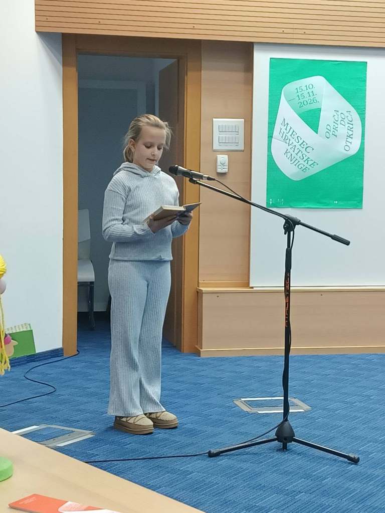 Djevojčica stoji za mikrofonom i čita iz knjige tijekom događanja u knjižnici.